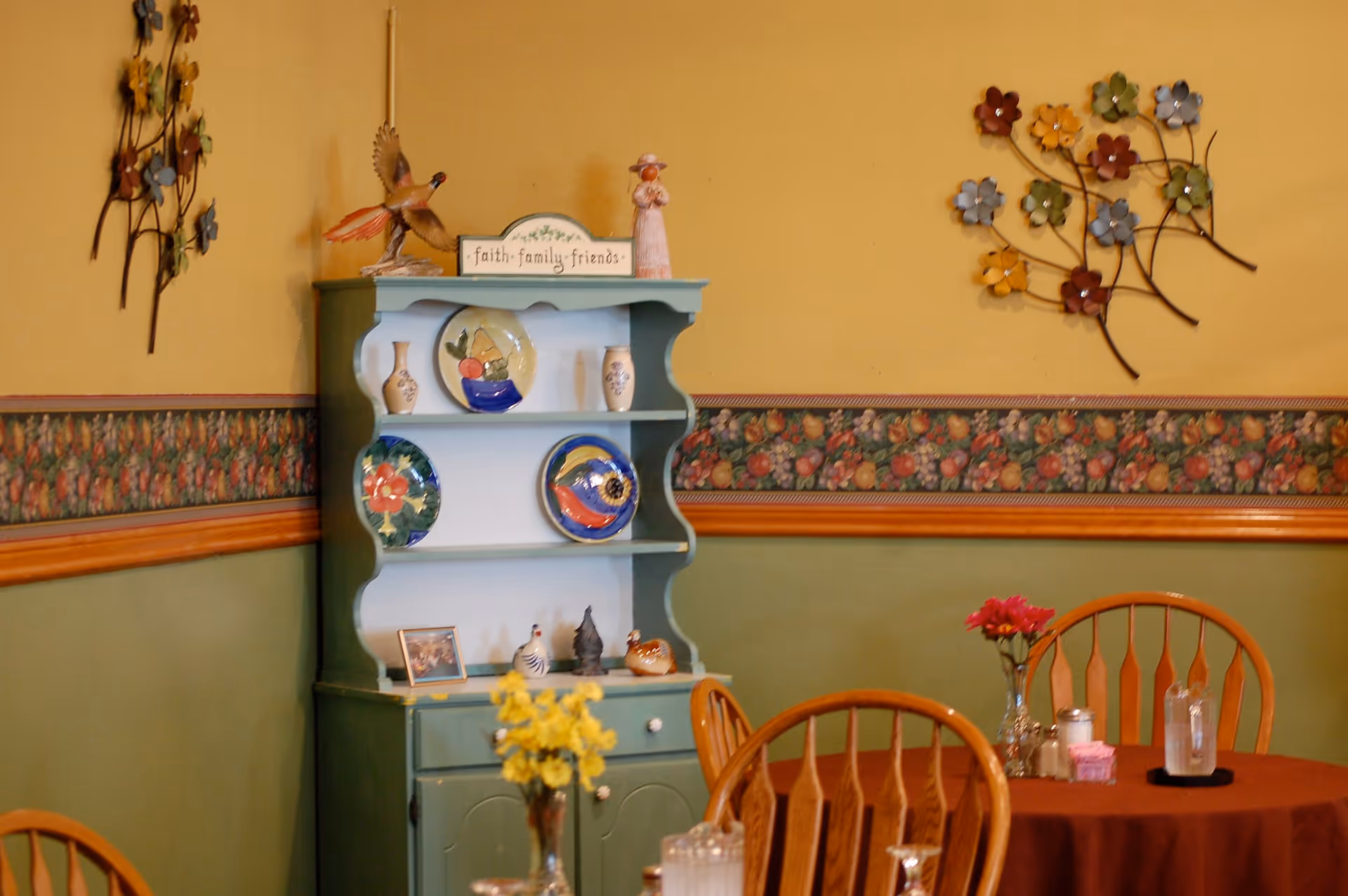 A cozy dining room corner with a green display hutch holding decorative plates and a table with wooden chairs and floral decor.