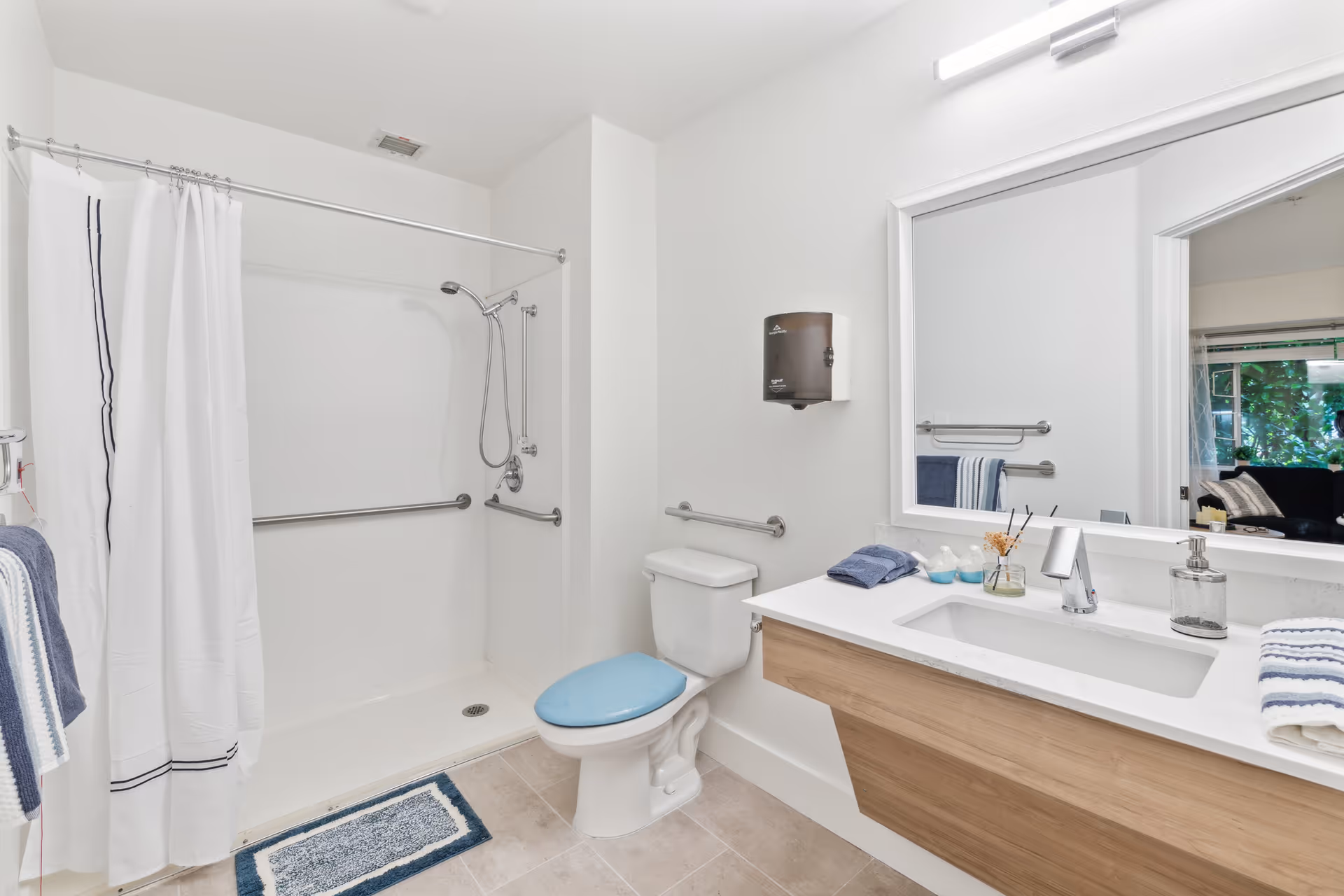 A clean, modern bathroom with a walk-in shower featuring grab bars and a white shower curtain. There is a toilet with a blue seat cover next to the shower. A wooden vanity with a white countertop and an under-mount sink is on the right side, with a large mirror above it. Towels and toiletries are neatly arranged on the countertop. The bathroom has light-colored tile flooring and white walls.