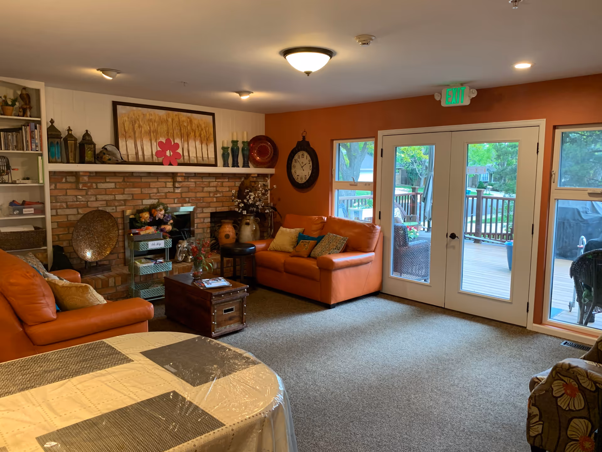 Cozy living room with two orange leather sofas, a brick fireplace with decorative items on the mantel, a wooden coffee table, and a round table covered with a plastic tablecloth in the foreground. French doors and windows provide a view of an outdoor deck with chairs and greenery.