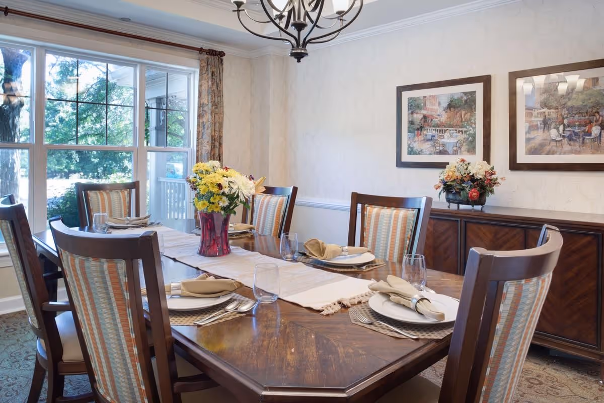 Sunlit dining room with a wooden table set for a meal, a floral centerpiece, striped upholstered chairs, and framed artwork on the wall.