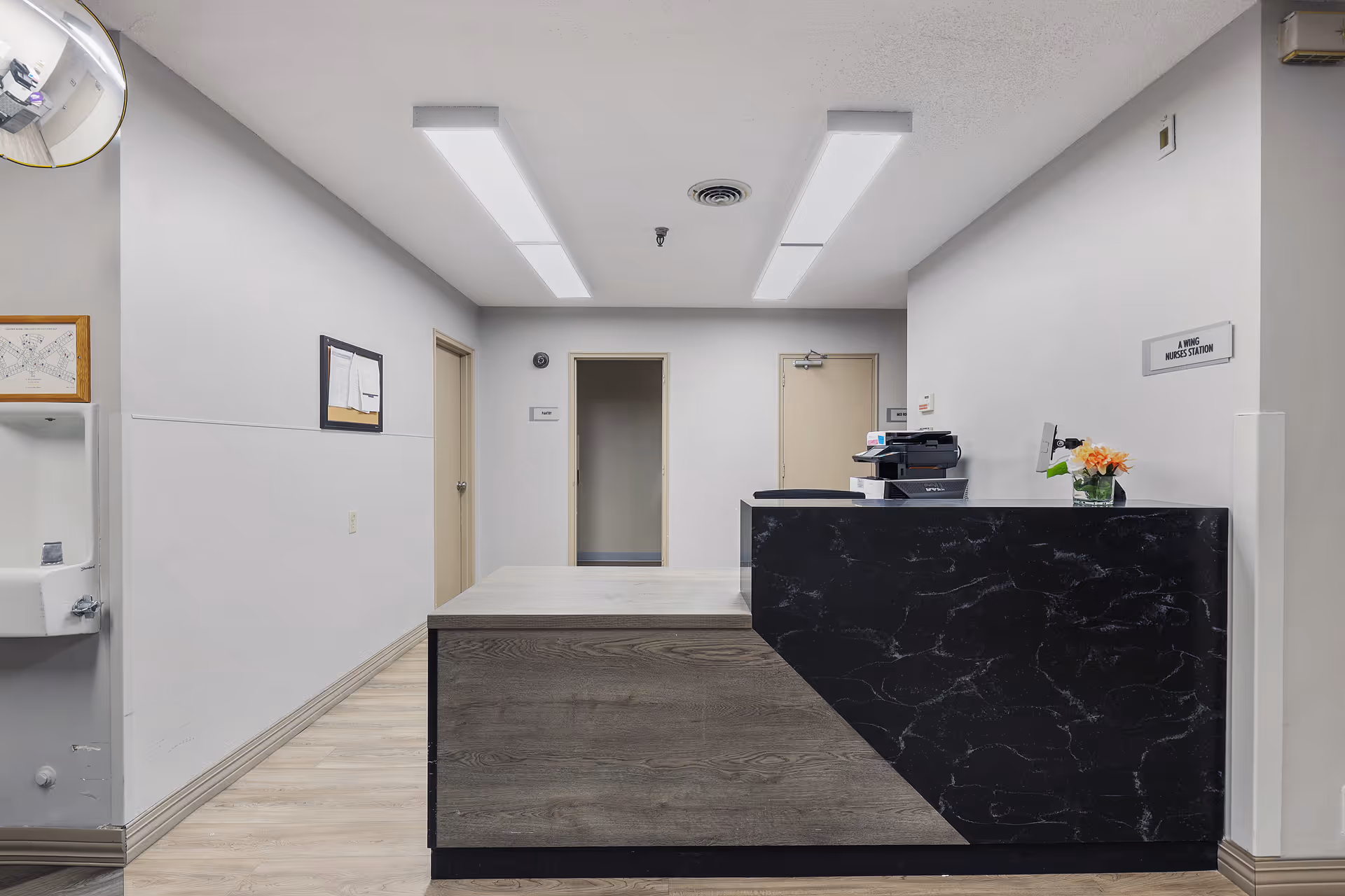 Interior view of a nurse station area in a healthcare facility with a black and wood reception desk, a printer, a small flower arrangement, and doors leading to other rooms. The walls are white, and there are fluorescent ceiling lights. A sign on the wall reads 'A Wing Nurses Station'.