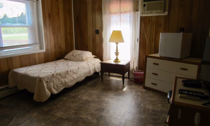 A small bedroom with wood-paneled walls featuring a single bed with a floral quilt and pillow, a wooden nightstand with a lamp, a window with blinds, a dresser with a small refrigerator on top, and an air conditioning unit mounted on the wall.
