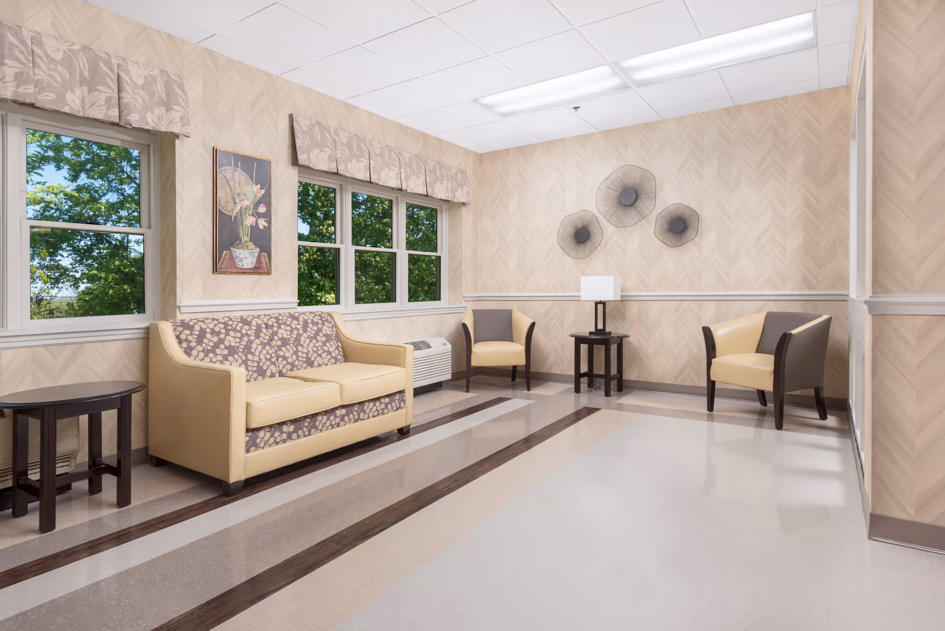 A well-lit seating area in a senior living facility with a beige and purple patterned sofa, two beige and brown armchairs, a small dark wooden side table, a lamp on a small table, and three decorative wall hangings. The room has large windows with beige valances showing green trees outside, beige patterned wallpaper, and a polished floor with brown and beige stripes.