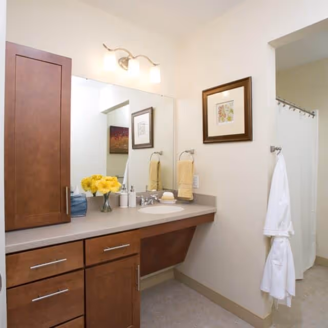 A clean and well-lit bathroom featuring a wooden vanity with a sink, a large mirror above it, and a vase of yellow flowers. There are two yellow hand towels hanging on a ring next to the sink, a framed picture on the wall, and a white bathrobe hanging on a hook near a shower with a white curtain.