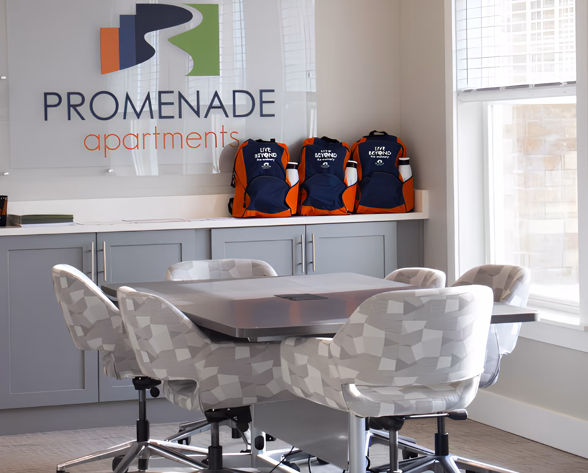 A modern meeting room with a rectangular table surrounded by six patterned swivel chairs. Behind the table is a counter with three orange and navy blue backpacks placed on it. Above the counter is a large sign that reads 'PROMENADE apartments'. The room has large windows allowing natural light to enter.