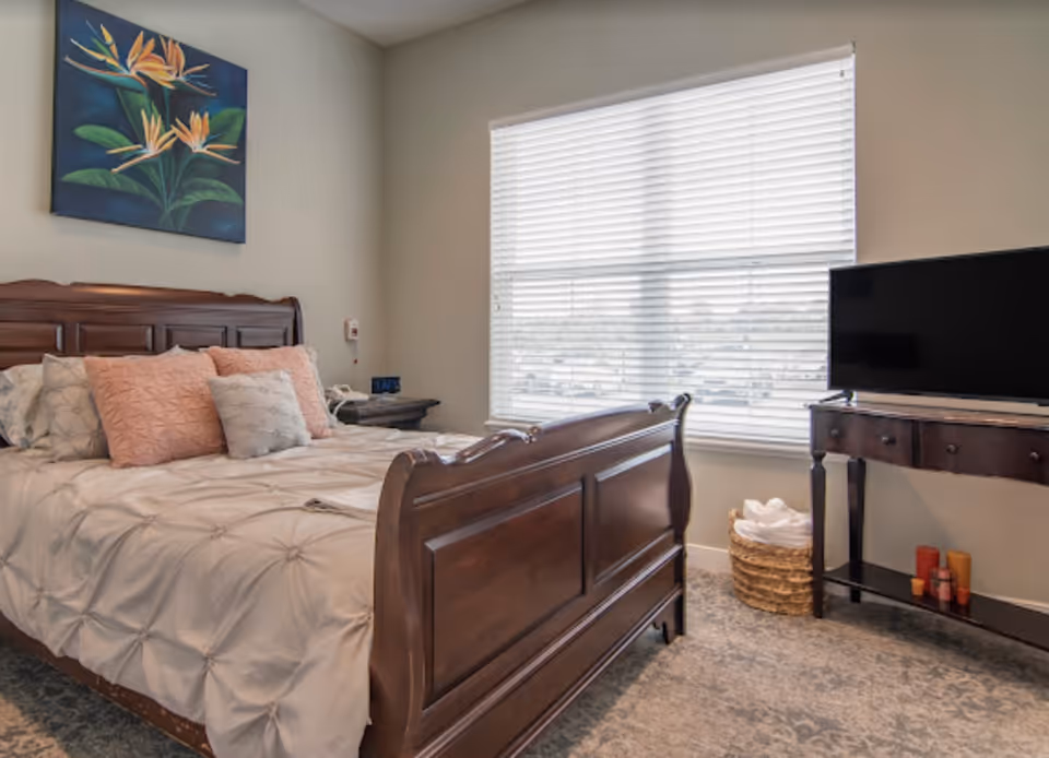 A bedroom with a wooden bed frame and a neatly made bed with white and pink pillows. There is a floral painting on the wall above the bed. Next to the bed is a nightstand with a phone and an alarm clock. A window with white blinds lets in natural light. Across from the bed is a dark wooden table with a flat-screen TV on top and a basket with towels underneath. The floor is carpeted.