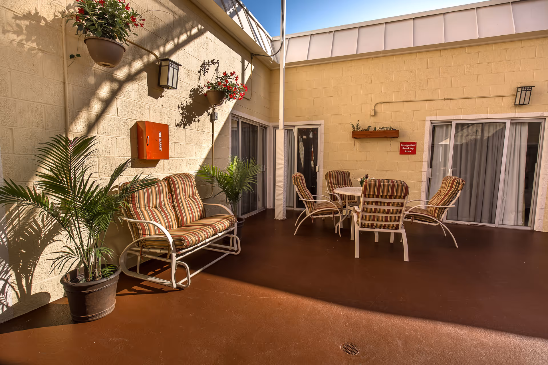 Outdoor patio area with striped cushioned seating including a loveseat and four chairs around a round table. The patio has potted plants, hanging flower pots, and a fire safety box mounted on a beige brick wall. Sliding glass doors lead inside the building.