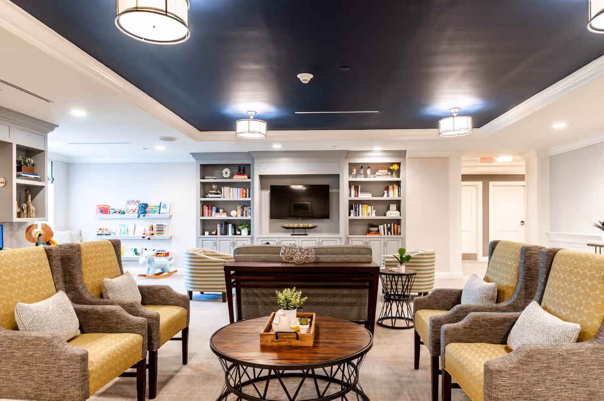 A bright and modern living room area in a senior living facility featuring comfortable armchairs with yellow and gray upholstery, a round wooden coffee table with decorative plants, a sofa, and built-in bookshelves flanking a wall-mounted flat-screen TV. The ceiling is painted dark blue with white crown molding and three ceiling lights. In the background, there is a small play area with children's toys and books on shelves.