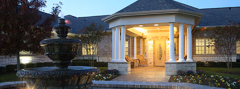 Evening view of the entrance to a brick building with a covered porch supported by white columns, warm lighting inside, a decorative water fountain in the foreground, and landscaped flower beds around the walkway.