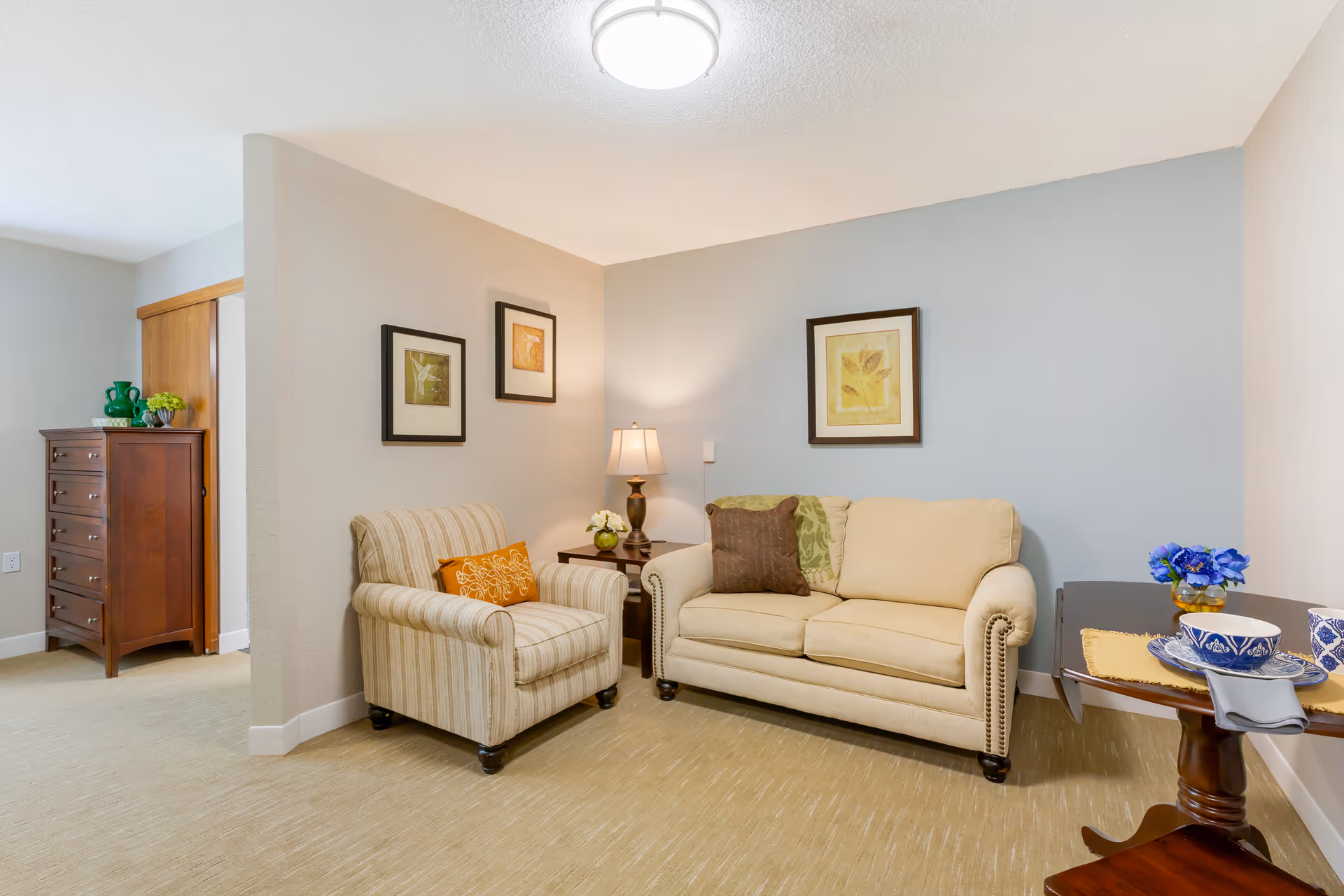 A cozy living room area with a beige sofa and a striped armchair, both adorned with decorative pillows. A wooden side table with a lamp and a small flower arrangement is placed between the sofa and armchair. On the right, a small round wooden table holds a blue and white bowl, a cup, and a small vase with blue flowers. The walls are painted in light beige and soft blue, decorated with framed botanical prints. In the background, a wooden chest of drawers with green decorative items is visible.