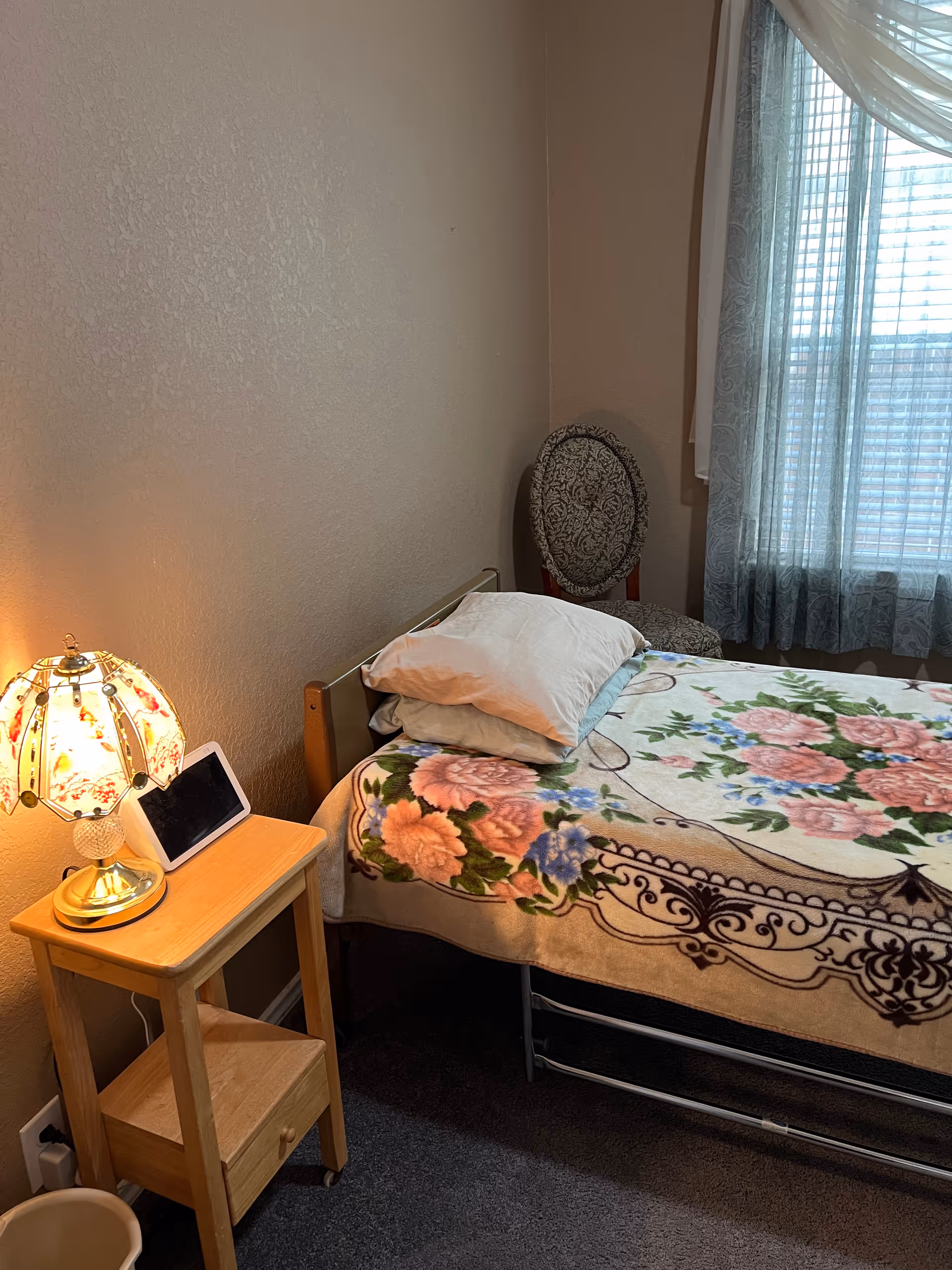 A small bedroom with a single bed covered by a floral blanket and two pillows. Next to the bed is a wooden nightstand with a decorative lamp and a digital device. A patterned chair is placed near a window with sheer curtains allowing light to enter the room.