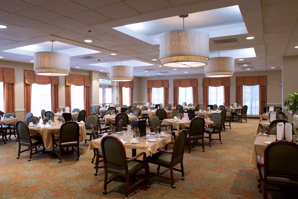 A spacious dining room with multiple round and rectangular tables covered with beige tablecloths. Each table is set with plates, glasses, and silverware, surrounded by green cushioned chairs with wooden frames. Large windows with brown curtains allow natural light to fill the room. The ceiling features recessed lighting and large, modern pendant lamps.