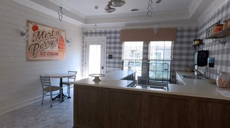 Interior view of a small ice cream shop area with a counter, a small round table with two chairs, and a sign on the wall that reads 'Mert & Perry's Ice Cream'. The walls have a mix of white shiplap and gray checkered wallpaper. There are pendant lights hanging from the ceiling and a window with a brown blind behind the counter.