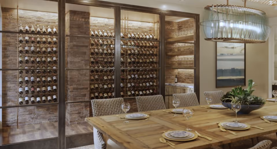 A dining room with a large wooden table set with plates, wine glasses, and gold-colored cutlery. There are six woven chairs around the table. Behind the table is a glass-enclosed wine rack filled with bottles, set against a brick wall. A modern light fixture hangs above the table, and a framed picture is visible on the wall.