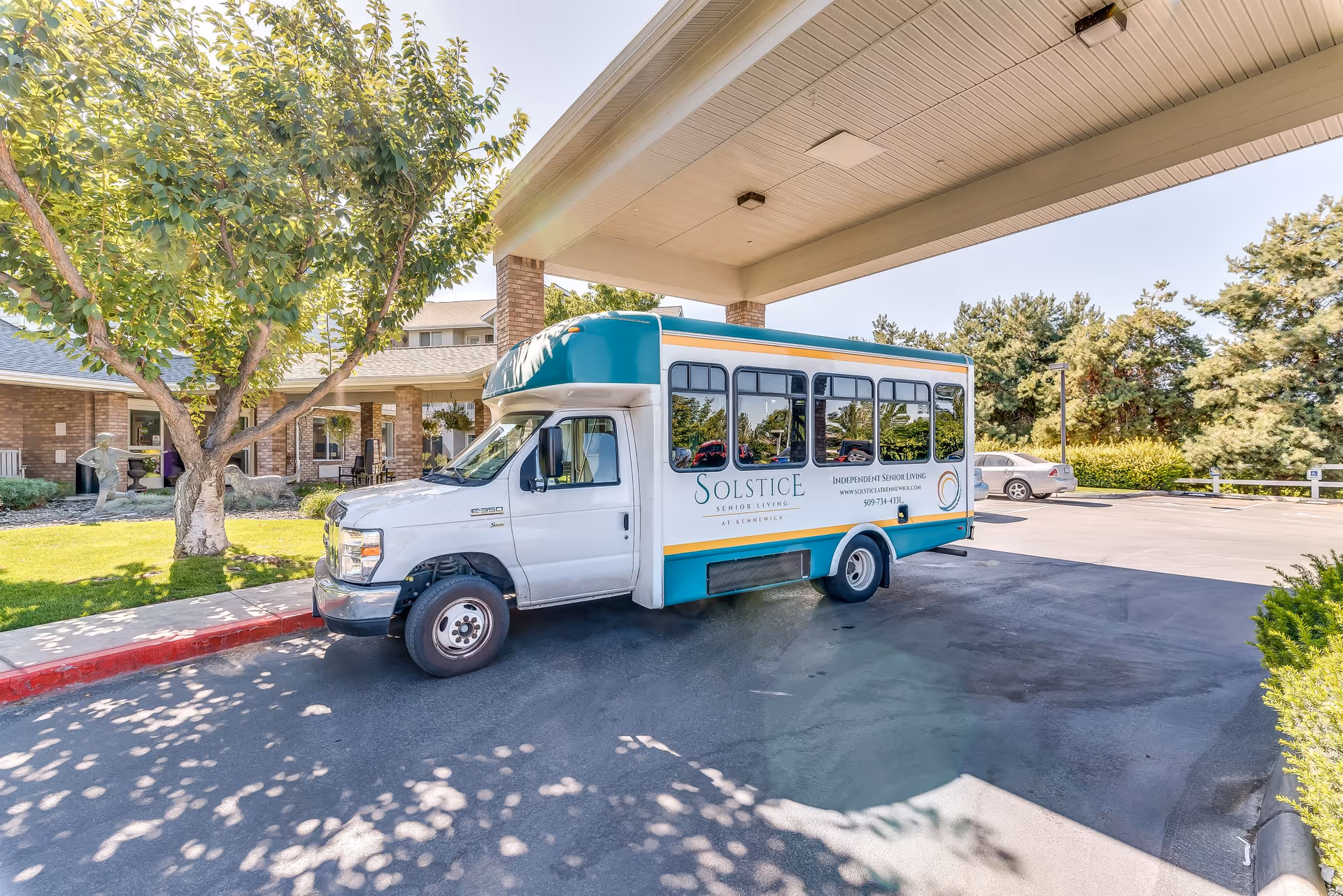 A white and teal shuttle bus with Solstice Senior Living branding parked under a covered entrance of a senior living facility. The building and surrounding greenery are visible in the background on a sunny day.