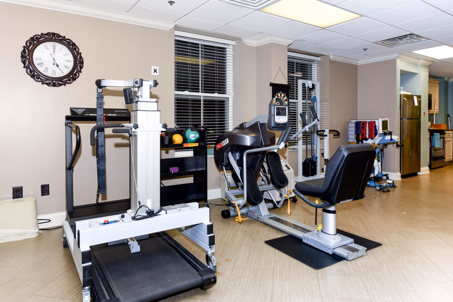 Interior view of a rehabilitation and healing center room featuring exercise equipment including a treadmill, a seated leg press machine, and various physical therapy tools. The room has beige walls, two windows with blinds, a decorative wall clock, and a kitchen area with a refrigerator and stove in the background.