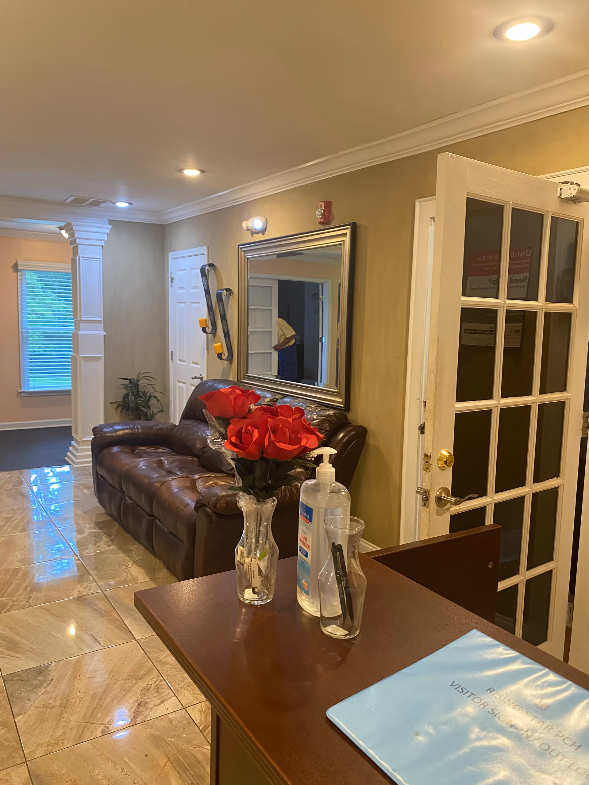 Interior view of a senior living facility reception area with a brown leather couch against a beige wall, a large mirror above the couch, a small table with a vase of red roses, hand sanitizer, and pens in a holder. There is a door with glass panels partially open, and a window with blinds in the background.