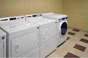 A laundry room with three white laundry machines: two top-loading washers and one front-loading dryer. The room has beige walls and a tiled floor with a pattern of beige and brown squares.