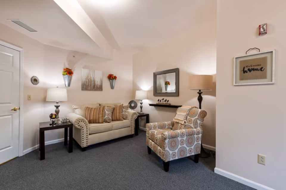 A cozy living room area with a beige sofa adorned with striped and patterned cushions, a patterned armchair with a throw blanket, two side tables each with a lamp, wall-mounted flower pots with red flowers, a framed mirror, and a framed 'Welcome Home' sign on the wall.