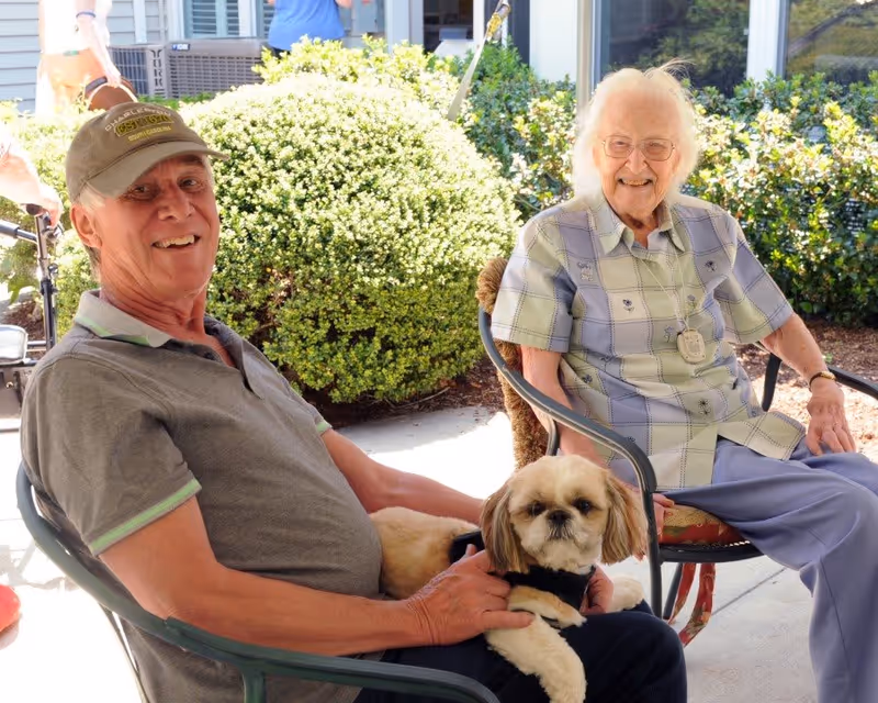 An elderly woman and an elderly man sitting outside on chairs. The man is holding a small dog on his lap. They are smiling and appear to be enjoying a sunny day in a garden or patio area with green bushes in the background.