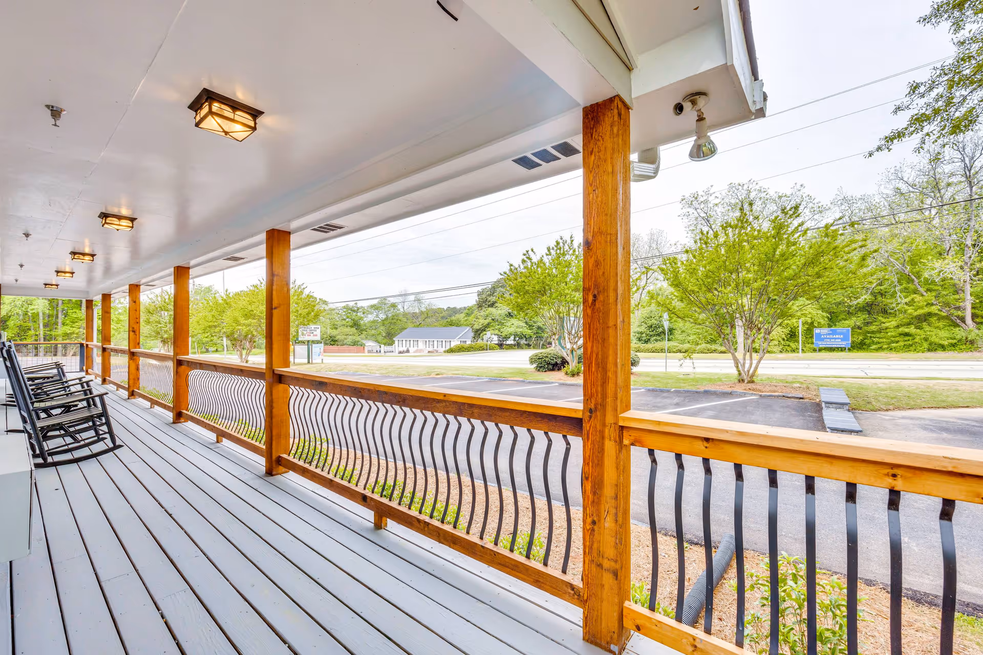 A covered outdoor porch with wooden posts and a railing featuring black metal balusters. Several black rocking chairs are lined up along the porch. The porch overlooks a parking area and a street with trees and greenery in the background under a cloudy sky.