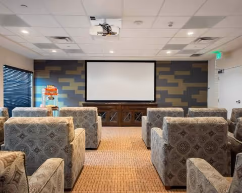 Upholstered armchairs arranged in rows facing a large projection screen in a small theater-style common room.