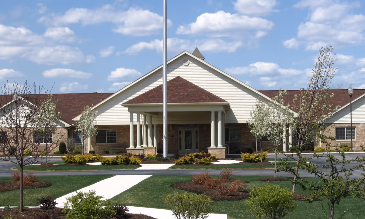 Front exterior view of Genoa Retirement Village - Senior Living Apartments building with a peaked roof, columns at the entrance, a flagpole, and landscaped grounds with small trees and shrubs under a partly cloudy sky.