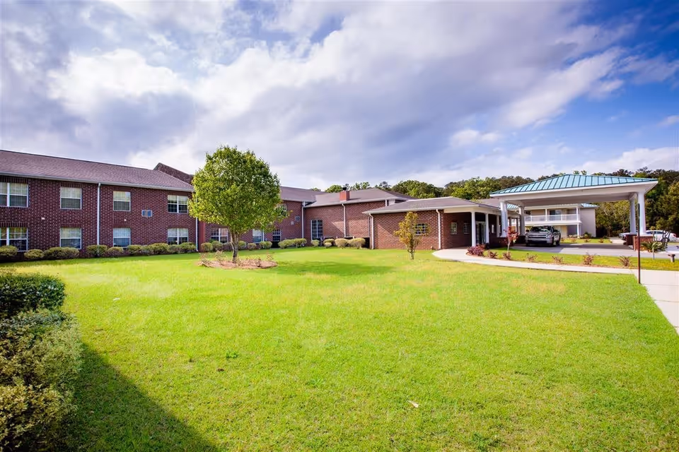 Exterior view of Hatton House facility showing a large green lawn with a few small trees, a brick building with multiple windows, and a covered entrance with a car parked underneath. The sky is partly cloudy.