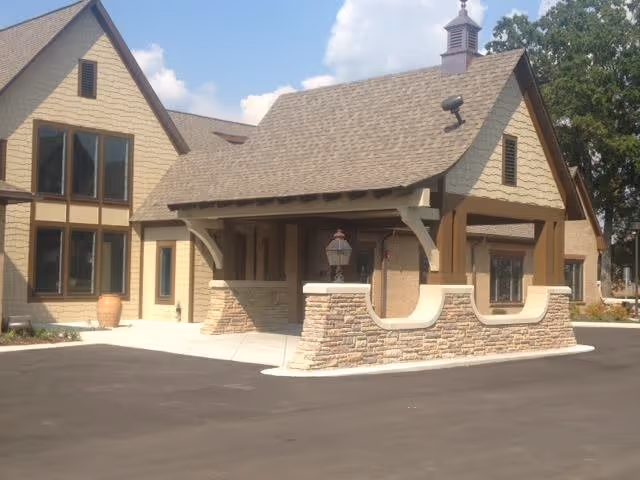 Exterior view of a building with beige siding and stone accents, featuring a covered entrance with a lamp post and large windows under a sloped roof with a small cupola on top. The sky is partly cloudy and there is a paved driveway in front.