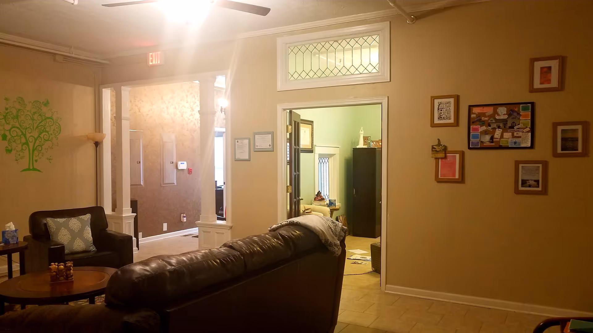 A cozy living room area with a dark leather sofa and a matching armchair with a patterned pillow. A round wooden coffee table is in front of the sofa. The walls are painted beige with a green tree decal on one wall and several framed pictures and notes on another. There is an open doorway leading to another room with a window and furniture visible. The room is softly lit with a ceiling fan light and a standing lamp in the corner.