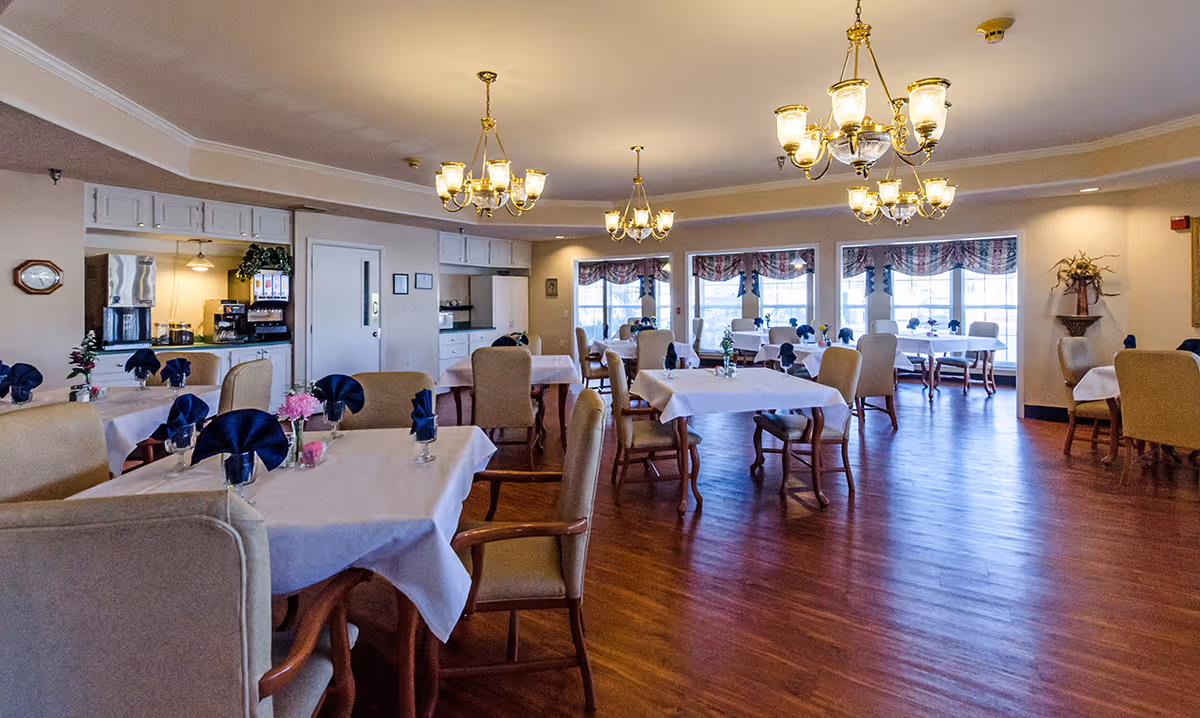 Spacious dining room with tables covered in white tablecloths, upholstered chairs, chandeliers, and large windows.