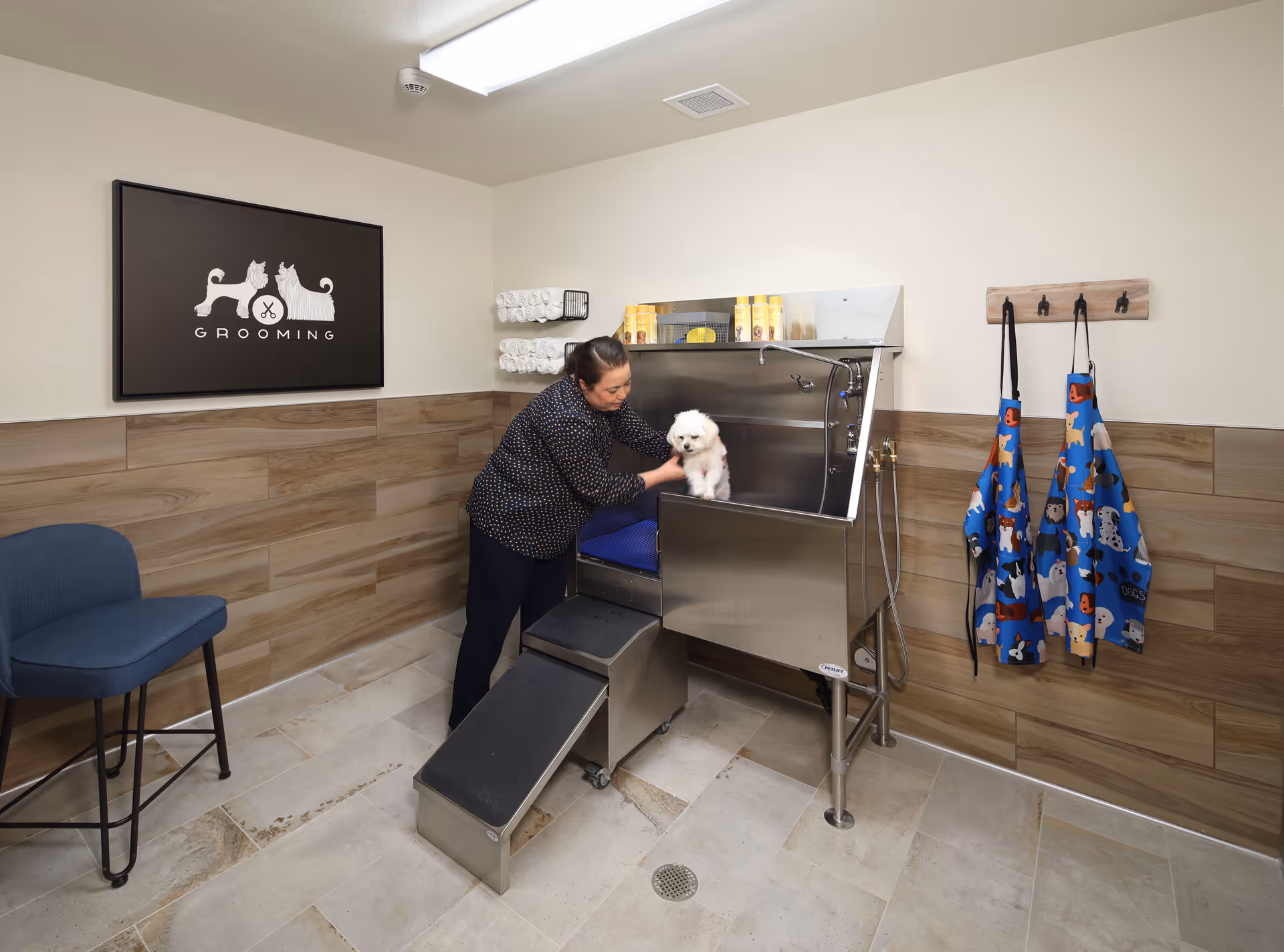 A woman is grooming a small white dog in a pet grooming station inside a room. The room has tiled floors and walls with wood paneling halfway up. There is a blue chair on the left, rolled white towels on a rack, grooming supplies on a shelf, and three aprons with dog prints hanging on hooks on the right wall. A framed sign with illustrations of a dog and a cat and the word 'GROOMING' is mounted on the wall.