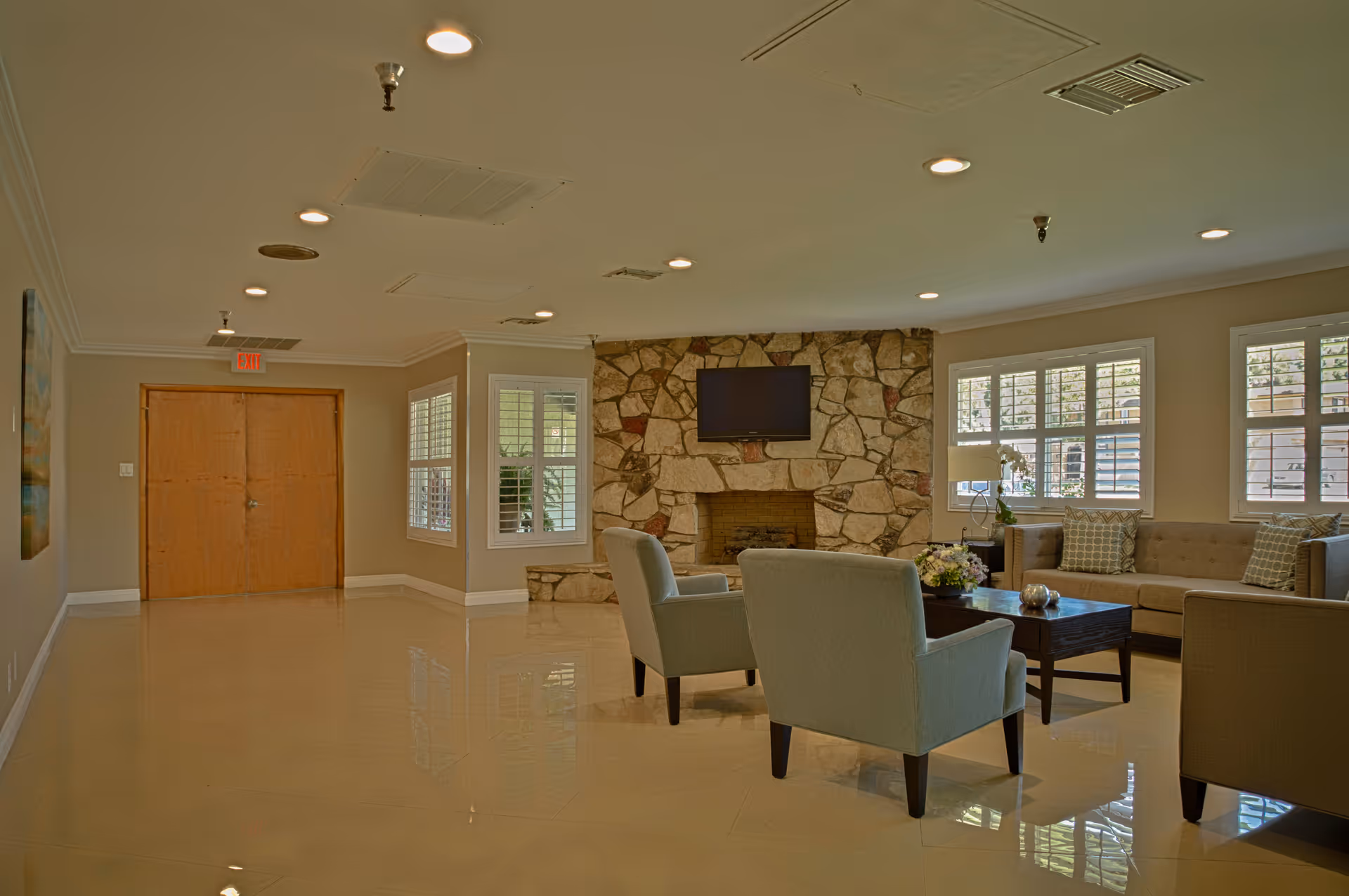 A spacious living room area with beige walls and shiny tiled floors. The room features a stone fireplace with a mounted flat-screen TV above it. There are several cushioned chairs and a sofa arranged around a dark wooden coffee table with a flower arrangement on top. Large windows with white shutters allow natural light to enter the room.