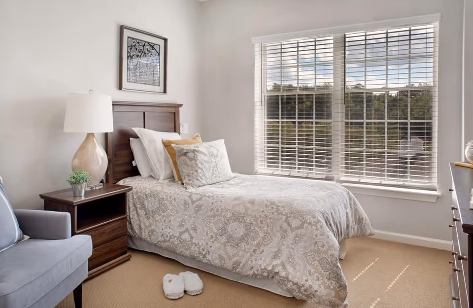 Bright bedroom with a single bed dressed in patterned bedding, a wooden nightstand and lamp, an armchair, a large window with blinds, and slippers on the carpet.