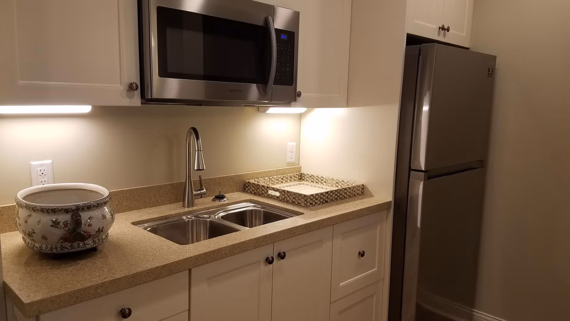 A modern kitchen area with white cabinets, a double stainless steel sink, a stainless steel microwave mounted above the sink, and a stainless steel refrigerator. The countertop is beige with a decorative bowl and a tray placed on it. Under-cabinet lighting illuminates the workspace.