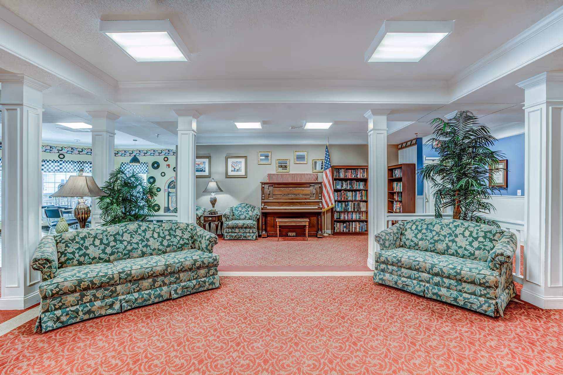 A spacious senior living facility common area with two floral patterned sofas facing each other on a red patterned carpet. The room features white pillars, a piano against the back wall, bookshelves filled with books, an American flag, framed pictures on the wall, and several large potted plants. The ceiling has recessed lighting panels.