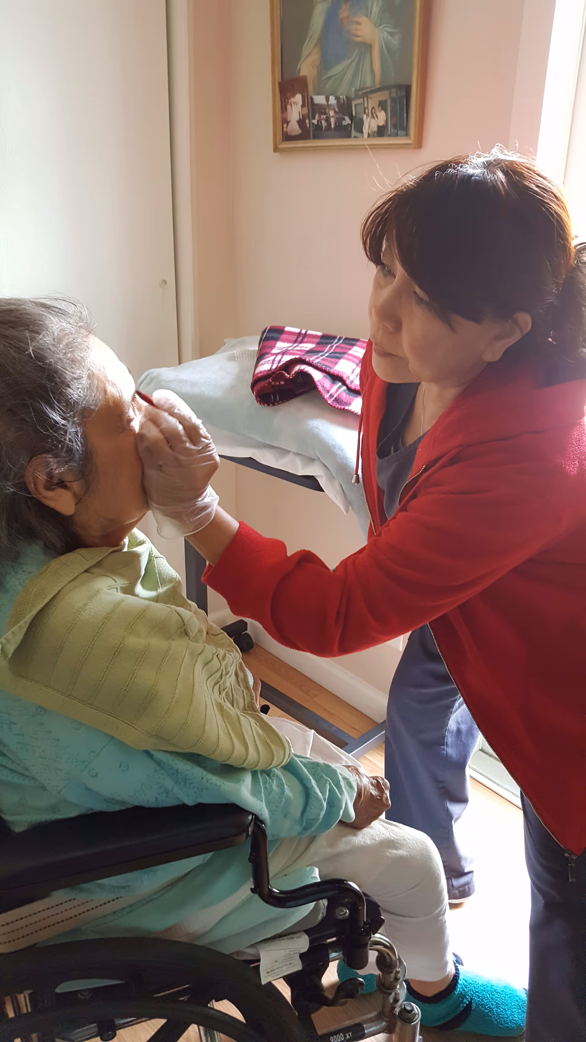A caregiver wearing a red jacket and gloves is gently wiping the face of an elderly woman seated in a wheelchair inside a room with light-colored walls and a framed picture on the wall.