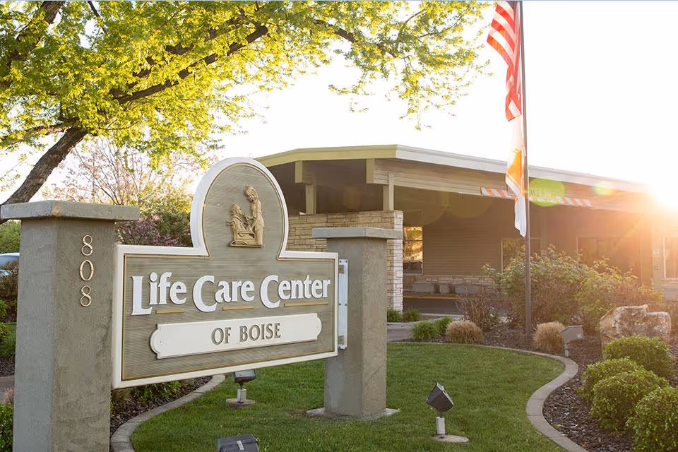 A wooden sign reading "Life Care Center of Boise" sits in landscaped grounds in front of the facility entrance with an American flag and sunlight in the background.
