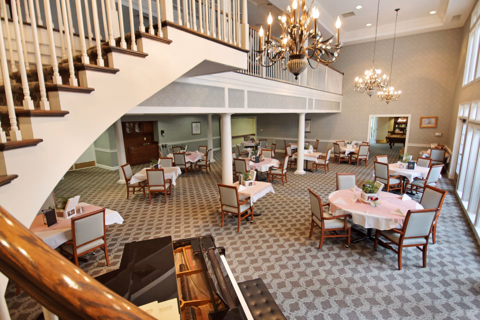 A spacious dining room with multiple round and square tables covered with white and pink tablecloths, surrounded by wooden chairs with light upholstery. The room features a patterned carpet, large windows allowing natural light, two elegant chandeliers hanging from the ceiling, and a grand piano in the foreground. A staircase with white railings and dark wooden handrails curves along the left side of the room.