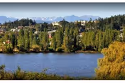 View across a calm lake toward a tree-lined shore with houses and distant mountains under a clear sky.