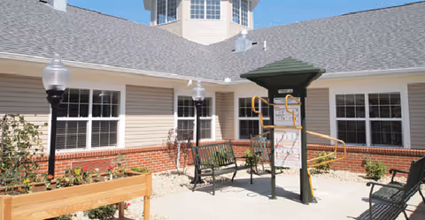 Outdoor courtyard area at The Willows at Willard featuring benches, a raised garden bed, instructional signage with a green roof, lamp posts, and a building with multiple windows and a gray shingled roof under a clear blue sky.