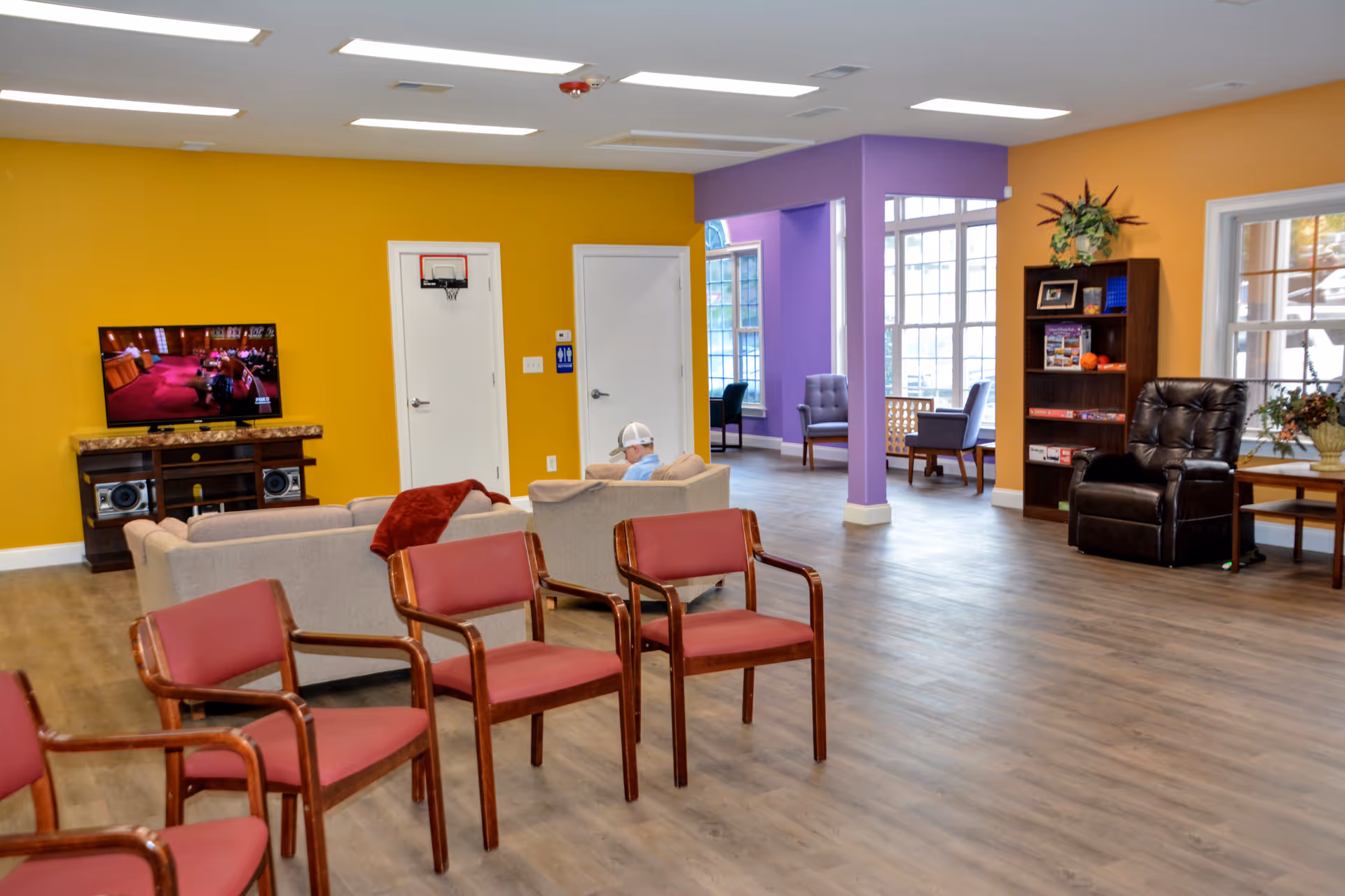 Bright communal lounge with chairs, sofas, a TV, bookshelves and large windows in a senior day center.