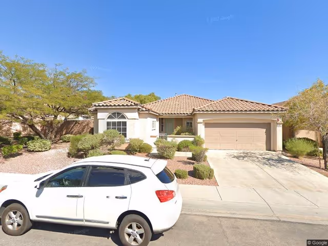 Single-story residential house with a tiled roof, beige exterior walls, a two-car garage, and desert landscaping including bushes and small trees. A white SUV is parked on the street in front of the house under a clear blue sky.