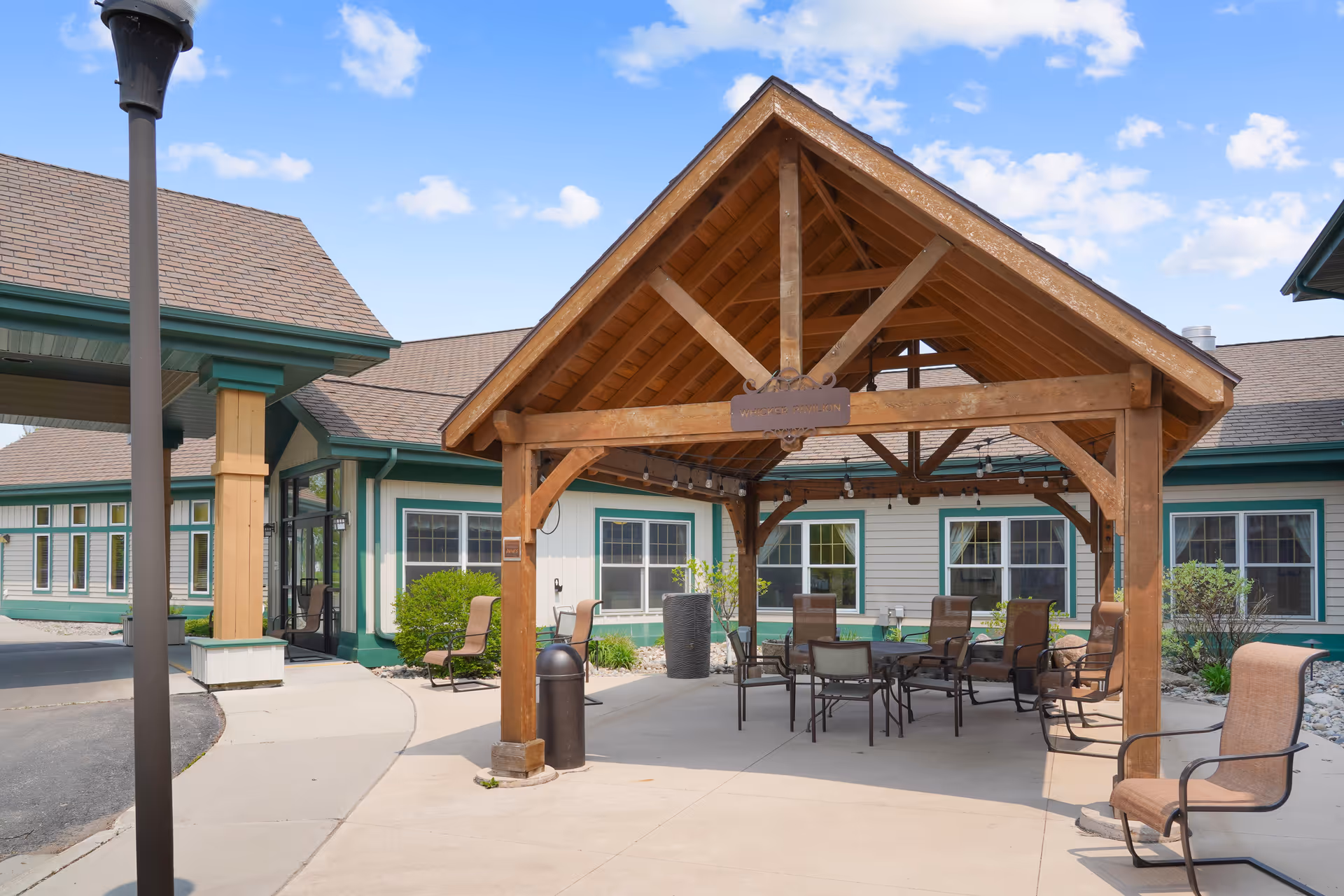 Wooden covered pavilion with tables and chairs in the outdoor courtyard in front of a single-story senior living building.