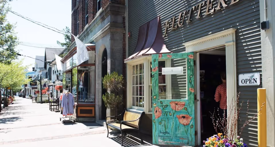 Sunlit street of storefronts showing an open shop with a painted green door, bench, clothing rack, and hanging flowers.