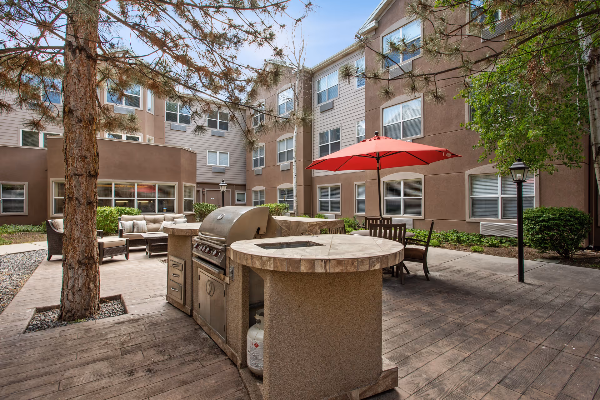 Outdoor courtyard area of a senior living facility with a built-in grill and countertop, a red patio umbrella shading a table with chairs, cushioned seating, trees, and surrounding multi-story building windows.