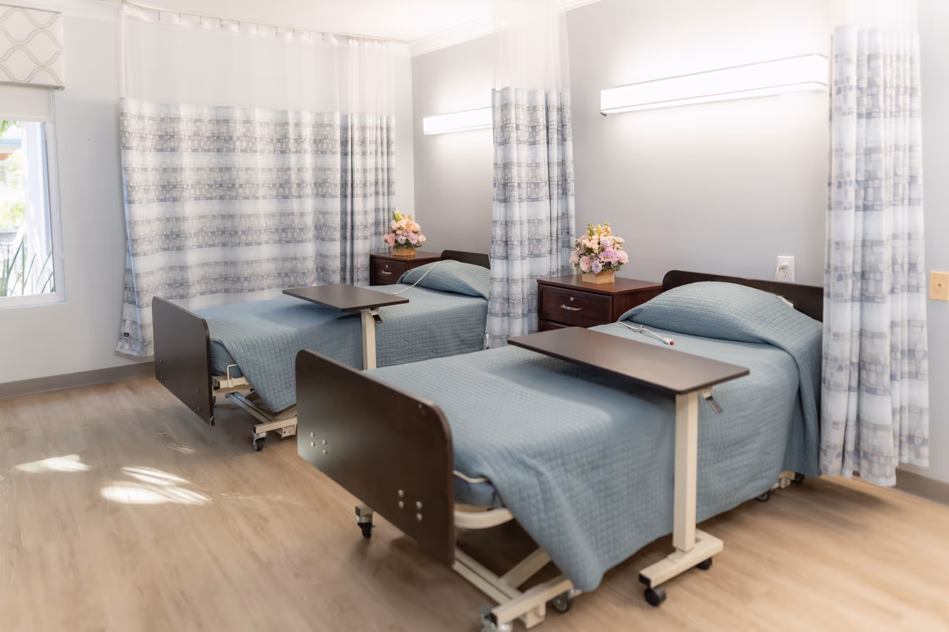 A shared senior care bedroom with two hospital-style beds, overbed tables, nightstands and floral arrangements.