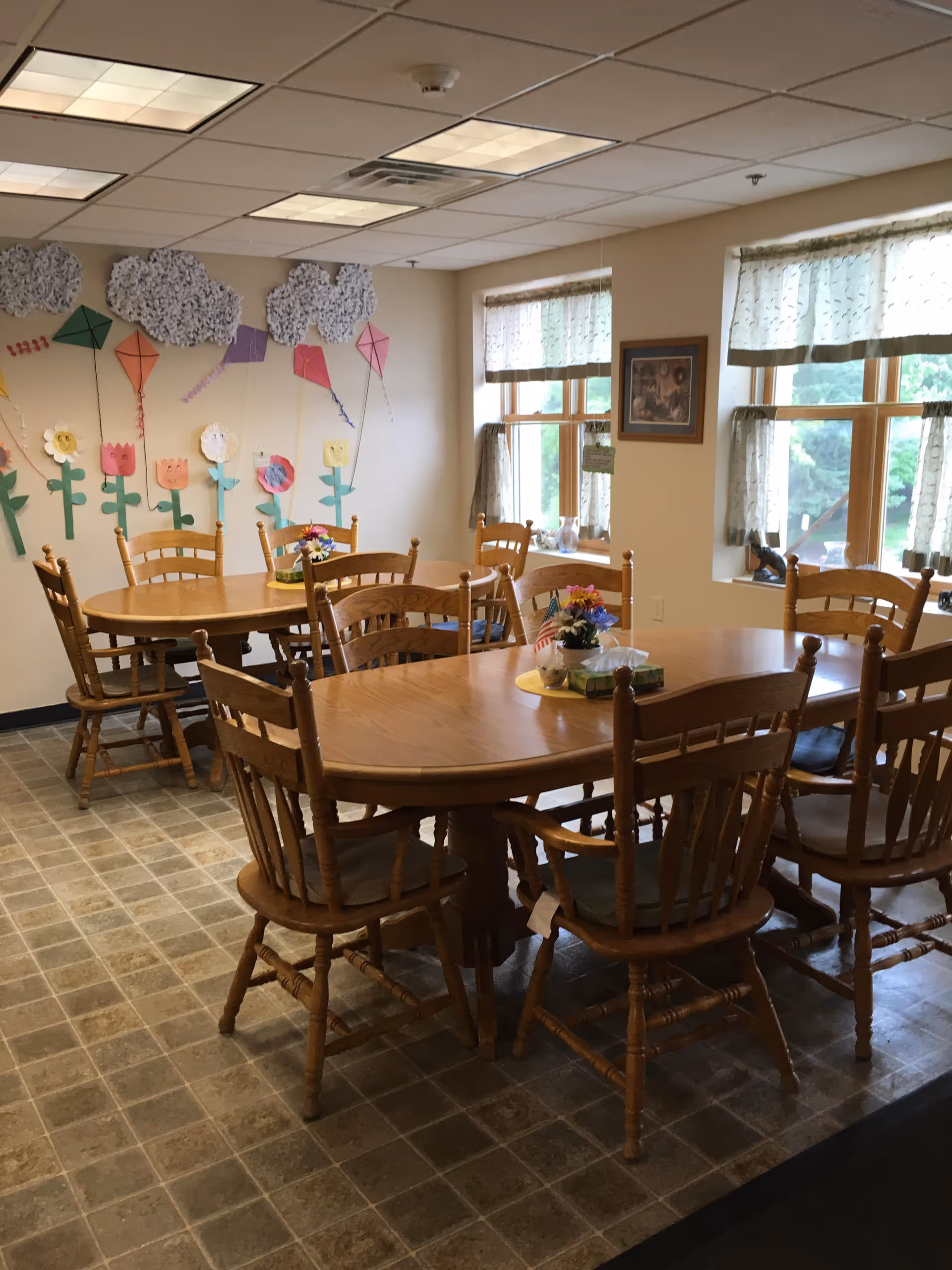 A dining room with two wooden tables surrounded by wooden chairs. The tables have small flower arrangements and tissue boxes on them. The room has two windows with light curtains, and the walls are decorated with colorful paper flowers and kites. The floor has a tiled pattern.