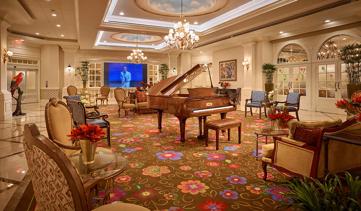 A spacious and elegant common area with a grand piano in the center, surrounded by various upholstered chairs and small tables with flower arrangements. The room features a colorful floral carpet, chandeliers hanging from a coffered ceiling with sky murals, large plants, and decorative wall sconces. A large screen on the far wall displays a person speaking, and there is a colorful parrot statue near the entrance.