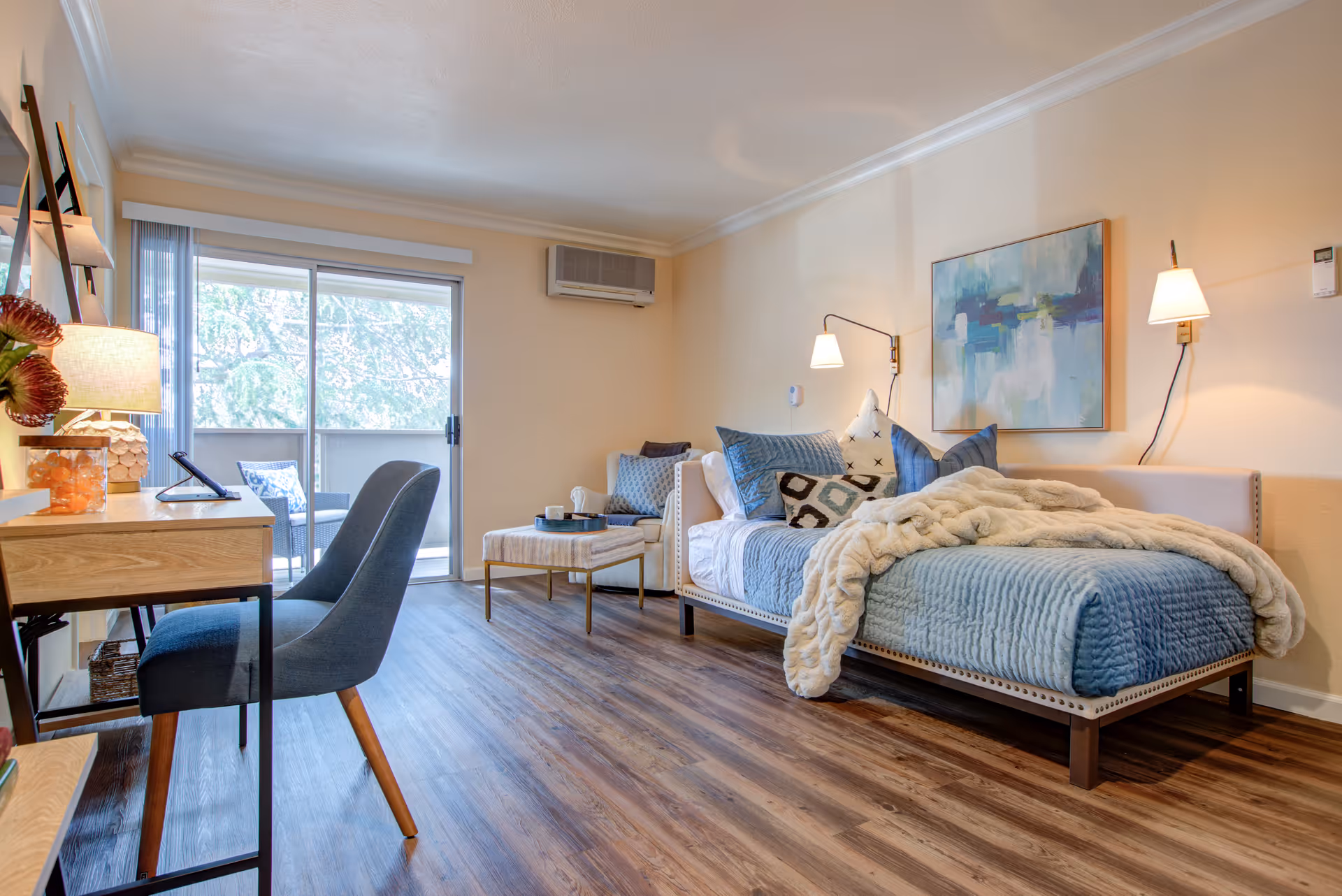 A cozy and well-lit bedroom with a single bed covered in blue bedding and a cream-colored throw blanket. The bed is adorned with multiple decorative pillows. Next to the bed is a small cushioned bench and a white armchair with a blue pillow. On the left side of the room, there is a wooden desk with a lamp, a chair, and some decorative items. The room has wood flooring and a large sliding glass door leading to a balcony with outdoor seating. The walls are painted a soft beige color, and there is a modern abstract painting hanging above the bed. Two wall-mounted lamps provide additional lighting.